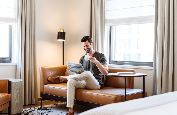 A man on a couch reading a paper and drinking a coffee in his suite at the Heathman Hotel in Portland, Oregon.